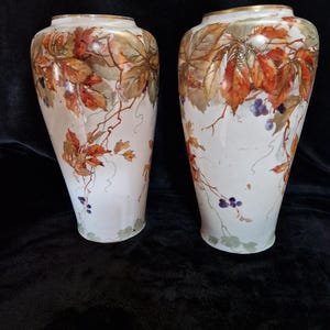 Puede incluir: Dos jarrones blancos con un diseño botánico. Los jarrones presentan un diseño pintado de hojas de otoño en tonos naranja, marrón y verde, junto con uvas moradas. Los bordes están adornados con oro. Los jarrones miden aproximadamente 30 cm de alto.