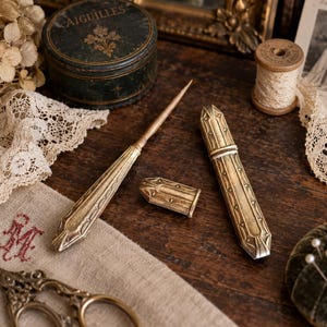 Pode incluir: Um estojo de agulhas em metal dourado com uma tampa removível a condizer e uma ferramenta de agulha longa e fina. O estojo apresenta um design alongado e facetado com padrões geométricos gravados ao longo da superfície metálica.