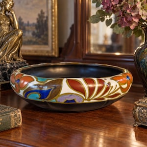Pode incluir: Uma taça de cerâmica larga e rasa com interior preto mate e uma borda arredondada decorada com um padrão floral vibrante pintado à mão em tons de laranja queimado, azul profundo, turquesa e lavanda suave sobre um fundo branco.