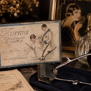 Puede incluir: Un set de cortapelos vintage Norma que incluye una caja de cartón rectangular con la ilustración de un barbero y un cliente, el texto "Norma Tondeuse à rasoir" y una cortapelos manual de metal con mango negro y cuchillas plateadas.