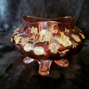 Peut inclure: Un bol en verre rouge rubis avec trois petits pieds. Le bol est orné de motifs floraux et de feuilles dorées. Le bol a un large dessus ouvert et un corps arrondi. Le verre a une finition brillante.