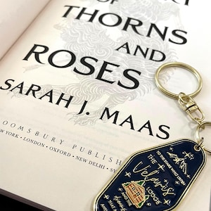May include: An open book with the title "A Court of Thorns and Roses" by Sarah J. Maas, next to a gold-colored keychain. The keychain is dark blue with gold trim and features the text "Velaris Lodge".