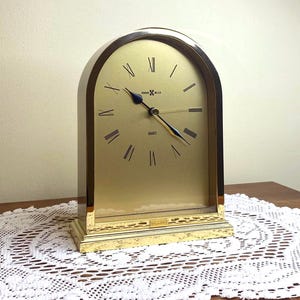 May include: A gold-toned Howard Miller Quartz clock with a curved top. The clock face is gold with black Roman numerals and hands. The clock is rectangular with a gold base and trim.