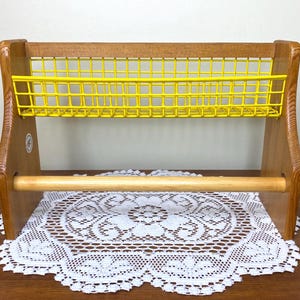 Op de afbeelding: Houten keukenorganizer met een felgele draadmand en een houten deuvel voor papieren handdoeken. De organizer is gemaakt van lichtgekleurd hout en heeft een eenvoudig, functioneel ontwerp. Onder de organizer ligt een decoratief wit kanten kleedje.