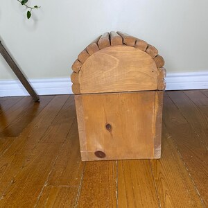 May include: A wooden chest with a domed lid, made from light-coloured wood. The lid is arched and has small, rounded wooden pieces along the top edge. The chest is rectangular with a visible knot on the front.