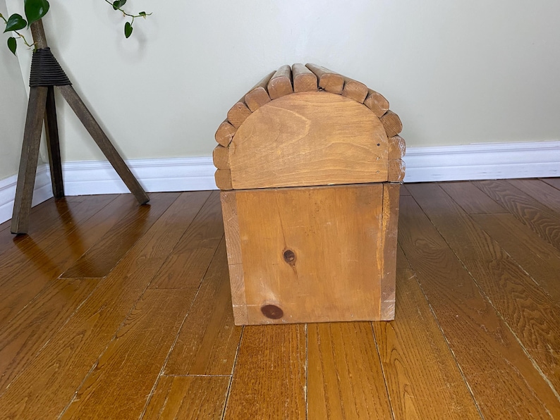 May include: A wooden chest with a domed lid, made from light-coloured wood. The lid is arched and has small, rounded wooden pieces along the top edge. The chest is rectangular with a visible knot on the front.
