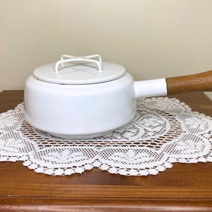 May include: White enamel saucepan with a lid and a wooden handle. The saucepan sits on a white lace doily on a wooden surface. The lid has a white handle. The handle is made of wood.