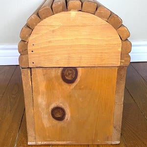 May include: A wooden storage chest with a rounded top, made from light-coloured wood. The top is constructed from rounded wooden pieces, and the front has two prominent knots. The chest has a rustic design.