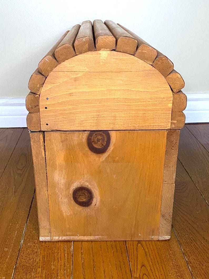May include: A wooden storage chest with a rounded top, made from light-coloured wood. The top is constructed from rounded wooden pieces, and the front has two prominent knots. The chest has a rustic design.
