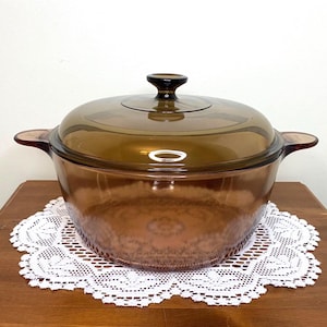 Può includere: Una pirofila in vetro marrone con coperchio abbinato. Il piatto ha due manici laterali e un pomello rotondo sul coperchio. La pirofila è appoggiata su una tovaglietta di pizzo bianco su una superficie di legno.