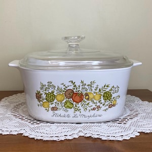 Pode incluir: Travessa branca com tampa de vidro transparente. A travessa apresenta um design botânico colorido de vegetais e ervas. O texto "L'Echalote La Marjolaine" está impresso na frente.