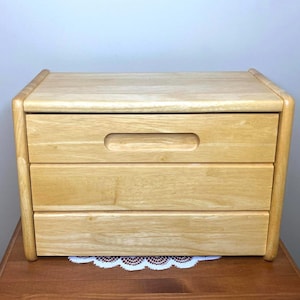 May include: A light wood bread bin with three drawers and a rounded top. The drawers have a simple, rectangular cutout for a handle.