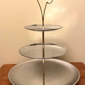 May include: A three-tiered silver serving stand with a gold handle. The stand has three round, mirrored plates.