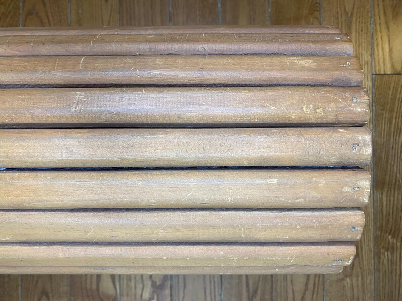 May include: Close-up of a light brown wooden bench with a slatted design. The bench has a weathered appearance with visible wood grain and minor imperfections, suggesting age and use. The design is simple and rustic.
