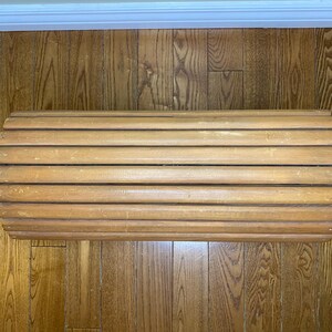 May include: A light brown wooden bench with a rounded shape. The bench is made of horizontal wooden slats, creating a ribbed texture. The bench is placed on a wooden floor.