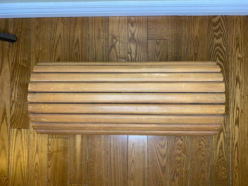 May include: A light brown wooden bench with a rounded shape. The bench is made of horizontal wooden slats, creating a ribbed texture. The bench is placed on a wooden floor.