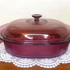 May include: An oval casserole dish with a lid, in a deep purple hue. The dish has a ribbed design on the sides and sits on a white lace doily. The lid has a small handle.