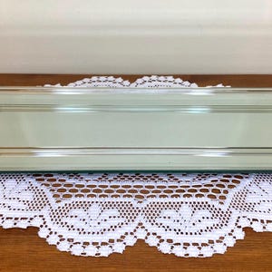 Op de afbeelding: Een rechthoekige spiegeltray met heldere acryl zijkanten en goudkleurige metalen accenten. De tray ligt op een witte kanten placemat en een houten oppervlak. De tray is een decoratief item.