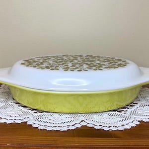 Peut inclure: Un plat à casserole Pyrex vintage avec un couvercle blanc orné d'un motif floral doré. Le plat est jaune et repose sur une nappe en dentelle blanche. Le plat a des poignées de chaque côté.