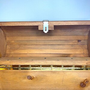 May include: Open wooden chest with a hinged lid. The chest is made of light brown wood with brass-coloured hinges and a metal latch. A metal chain is attached to the lid to prevent it from opening too far. The interior is visible.