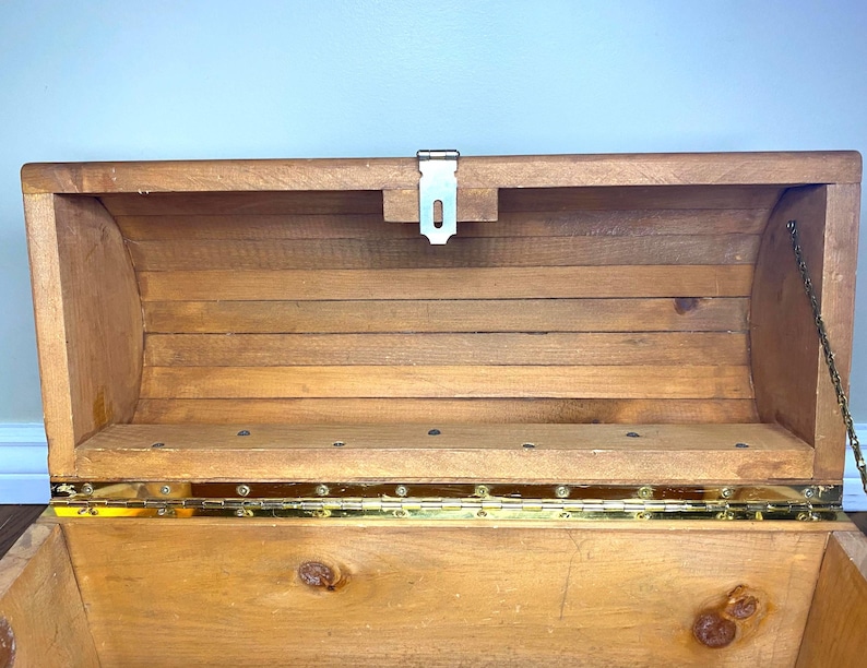 May include: Open wooden chest with a hinged lid. The chest is made of light brown wood with brass-coloured hinges and a metal latch. A metal chain is attached to the lid to prevent it from opening too far. The interior is visible.