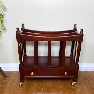 May include: A dark brown wooden magazine rack featuring a rectangular frame with four turned corner posts, a central divider, and a single bottom drawer with two small round gold-toned knobs. The unit is supported by four small metal casters.