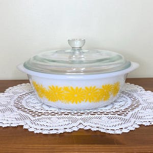 Peut inclure: Plat à casserole blanc avec un couvercle en verre transparent et une petite poignée. Le plat est décoré d'un motif de marguerites jaunes à l'extérieur. Le plat est posé sur un napperon en dentelle blanche.