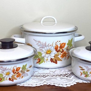 Op de afbeelding: Een set van drie vintage witte emaille kookpotten met deksels. De potten hebben een bloemenpatroon in oranje, geel en groen. Ze hebben bruine handvatten en zwarte knoppen. De set bevat een grote pot en twee kleinere.