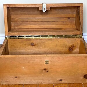 May include: Open wooden chest with a hinged lid, metal clasp, and brass hinges. The chest is made of light-coloured wood with visible knots and grain. A metal chain is attached to the lid. The chest is empty and sits on a wooden floor.
