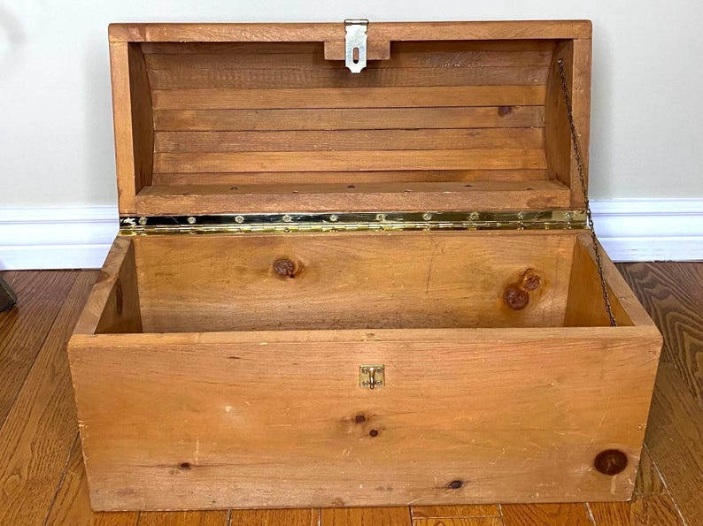 May include: Open wooden chest with a hinged lid, metal clasp, and brass hinges. The chest is made of light-coloured wood with visible knots and grain. A metal chain is attached to the lid. The chest is empty and sits on a wooden floor.