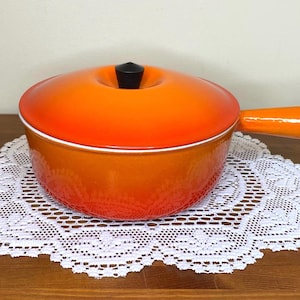 Op de afbeelding: Een oranje geëmailleerde gietijzeren braadpan met bijpassende deksel en handvat. De deksel heeft een zwarte knop. De pot staat op een wit kanten kleedje op een houten oppervlak. De pot heeft een diameter van ongeveer 25 cm.