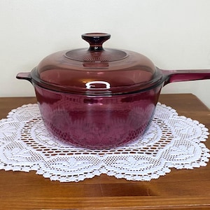 Op de afbeelding: Een vintage, dieppaarse glazen steelpan met bijpassende deksel. De steelpan heeft een lange handgreep en een ronde body. Hij staat op een wit kanten kleedje op een houten oppervlak. De deksel heeft een decoratief patroon.