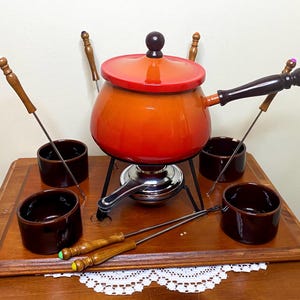Puede incluir: Un juego de fondue vintage con una olla naranja, tapa roja y soporte negro. Incluye cuencos de cerámica marrón y tenedores de fondue con mangos de madera. El juego se exhibe en una bandeja de madera con un tapete de encaje.