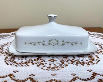 Könnte beinhalten: Eine weiße Butterdose aus Keramik mit rechteckigem Boden und passendem Deckel mit zentralem Knauf. Die Außenseite des Deckels ist mit einem zarten grün-weißen Blumenrankenmuster verziert und mit einem dünnen silbernen Metallrand versehen.