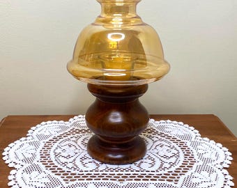 May include: A vintage candle lantern with a wooden base and a golden-amber glass shade. The base is a dark brown, turned wood design. The lantern sits on a white lace doily on a wooden surface. The overall aesthetic is a warm, rustic style.