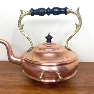 May include: A copper teapot with a brass handle and a black wooden knob. The teapot has a round body and a spout on the side. The handle is ornate and has a decorative design.