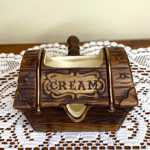 May include: A brown ceramic creamer in the shape of a barrel, with the word "CREAM" in a decorative font. The creamer has a handle and gold-colored accents. The creamer is sitting on a white lace tablecloth.