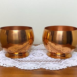 Peut inclure: Une paire de bols en cuivre poli avec une forme arrondie et une large base circulaire. Chaque bol présente un motif de dentelle gravé décoratif enveloppant l'extérieur inférieur, créant un contraste entre la finition métallique lisse.