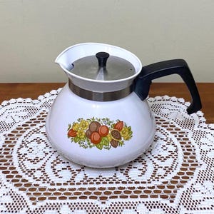 Könnte beinhalten: Weiße Emaille-Teekanne mit schwarzem Griff und Deckel. Die Teekanne hat ein Obst- und Blumendesign in Orange-, Braun- und Gelbtönen. Die Teekanne steht auf einer weißen Spitzendecke.