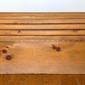 May include: A wooden storage chest with a hinged lid. The lid is made of several rounded wooden slats. The chest is made of light-coloured wood with visible knots and a brass hinge. The chest sits on a wooden floor.