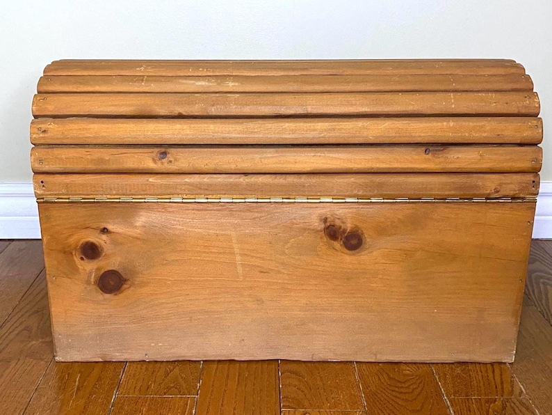 May include: A wooden storage chest with a hinged lid. The lid is made of several rounded wooden slats. The chest is made of light-coloured wood with visible knots and a brass hinge. The chest sits on a wooden floor.