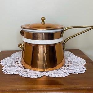 May include: A copper double boiler with a polished finish, featuring a white enamel rim and a matching lid with a small brass knob. The pot has two brass handles, one long and one short, and a cylindrical body with a flat base.