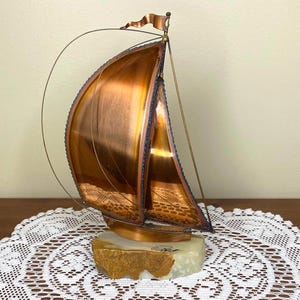 May include: A copper sailboat sculpture with a flag on top. The sails are a shiny copper colour, and the base is a light-coloured stone. The sculpture has a wire that arcs from the top of the mast to the back of the boat.