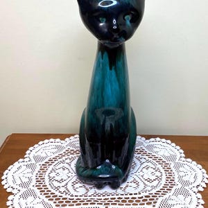 May include: A ceramic cat ornament with a teal and dark blue glaze. The cat is seated with pointed ears and a long body. The ornament is displayed on a wooden surface with a white lace doily. The glaze has a glossy finish.