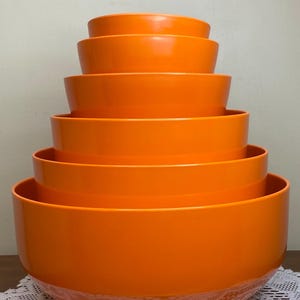 Könnte beinhalten: Ein Satz von sechs leuchtend orangefarbenen Rührschüsseln unterschiedlicher Größe, die übereinander gestapelt sind. Die Schüsseln bestehen aus glänzendem Material und stehen auf einer weißen Spitzendecke. Die Schüsseln haben eine leuchtende, einfarbige Farbe.