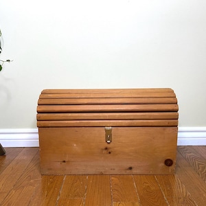 May include: A wooden chest with a curved, slatted top and a rectangular body. The chest is made of light-coloured wood and features a brass-coloured latch. The chest is sitting on a wooden floor.