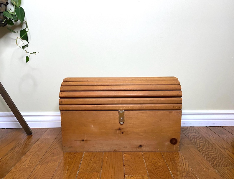 May include: A wooden chest with a curved, slatted top and a rectangular body. The chest is made of light-coloured wood and features a brass-coloured latch. The chest is sitting on a wooden floor.