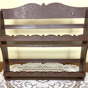 May include: A brown wooden spice rack with three shelves and a decorative scalloped edge. The rack has a textured wood grain appearance and is designed to be mounted on a wall. The top of the rack has a decorative cutout design.