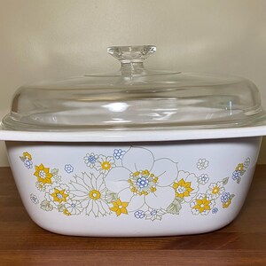 May include: White casserole dish with a floral design in yellow, blue, and green. The dish has a clear glass lid with a knob.