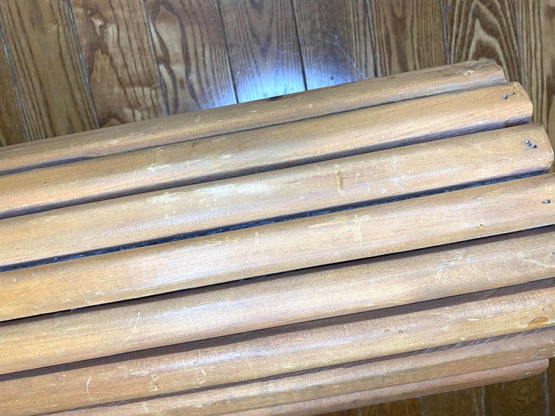 May include: A close-up of several light brown wooden dowels arranged in parallel. The dowels are cylindrical and appear to be made of natural wood. The wood grain is visible, and there are small holes drilled into the ends of some of the dowels.